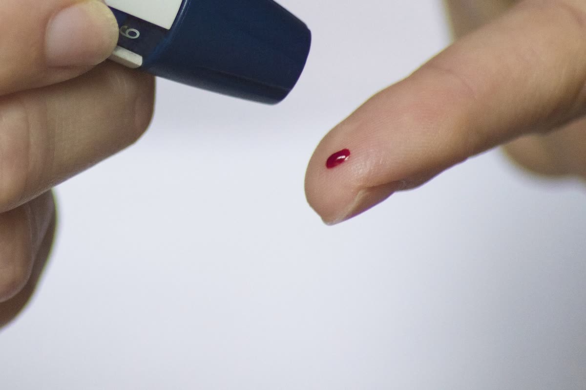 A lancing device held against a fingertip, with a bead of blood on the pad, on a plain white background