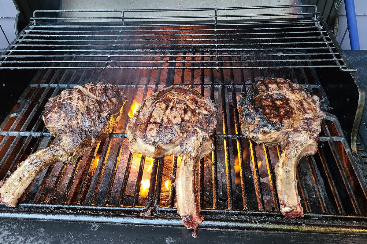Three tomahawk steaks searing on a gas grill, the trimmed bones pointing towards the camera