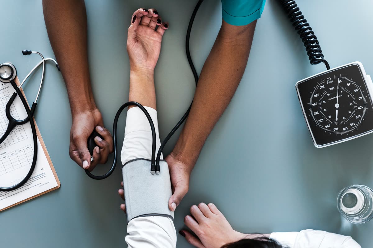 Hands placing a grey blood pressure cuff on a patient's arm beside a stethoscope and an aneroid gauge on a blue clinic desk